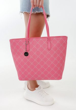 Osoba trzymająca dużą różową torbę na ramię z białym geometrycznym wzorem, ubraną w postrzępioną dżinsową spódnicę i białe sneakersy na platformie, na białym tle.