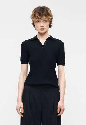 Person med kort, bølget hår iført en sort kortærmet skjorte med krave og sorte bukser med fine striber, stående mod en ensfarvet lys baggrund.