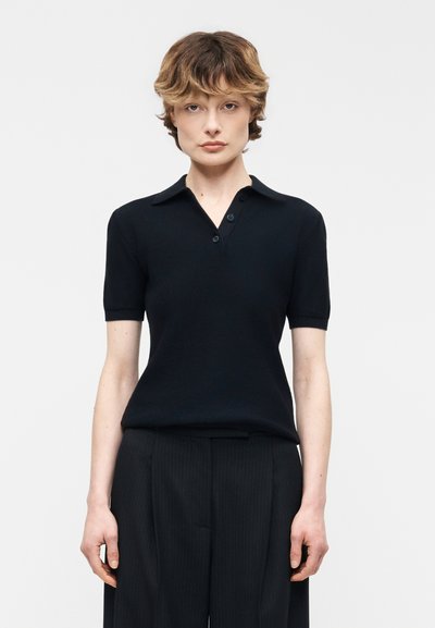 Personne aux cheveux courts et ondulés portant une chemise noire à manches courtes avec col et un pantalon noir à rayures fines, debout devant un fond clair uni.