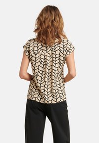 Short-sleeve blouse in beige with a black leaf pattern. The fabric has a smooth texture, featuring a relaxed fit and a back yoke detail.