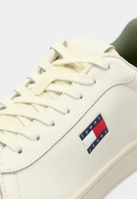 White synthetic sneaker with cream laces, featuring a logo badge in red, white, and navy. Rounded toe and flat rubber sole.