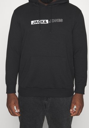 Czarna bluza z kapturem z przednią kieszenią i białym logo "JACK & JONES" na piersi, noszona przez osobę o ciemnej karnacji i ciemnych spodniach.