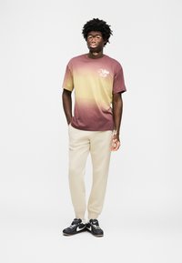 Μπλούζα με διαβάθμιση χρώματος από μαρόν σε κίτρινο, με μικρό λογότυπο Nike. Συνδυασμένη με κρεμ φόρμες και μαύρα αθλητικά παπούτσια. Ομαλή υφή υφάσματος.