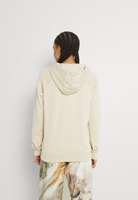 Beige huvtröja med raglanärmar, mjukt tyg och en lätt avslappnad passform, kombinerad med mönstrade flerfärgade byxor.