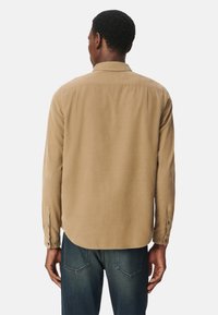 Koszula z długim rękawem w jasnobrązowym materiale o gładkiej teksturze, z klasycznym kołnierzykiem i zapięciem na guziki, noszona z niebieskimi dżinsami.