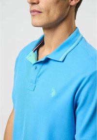 Šviesiai mėlynas polo marškinėliai su dviem sagomis, kontrastinga mėtų žalumo apykakle ir subtiliu siuvinėtu logotipu kairiajame krūtinės pusėje.