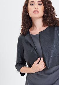 Giacca cropped in eco-pelle nera con collo rotondo e maniche a tre quarti, abbinata a una maglia grigia. Texture liscia, dettagli minimi.