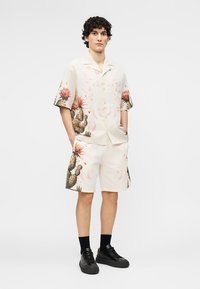 Young man wearing cream matching short-sleeve shirt and shorts with cactus and flower print, black socks, and black sneakers, hands in pockets.