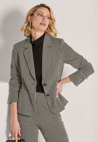 Blonde Frau trägt einen schwarzen Rollkragenpullover sowie einen Blazer und eine Hose im Schachbrettmuster, hält eine schwarze Handtasche und schaut seitlich vor einem schlichten Hintergrund.