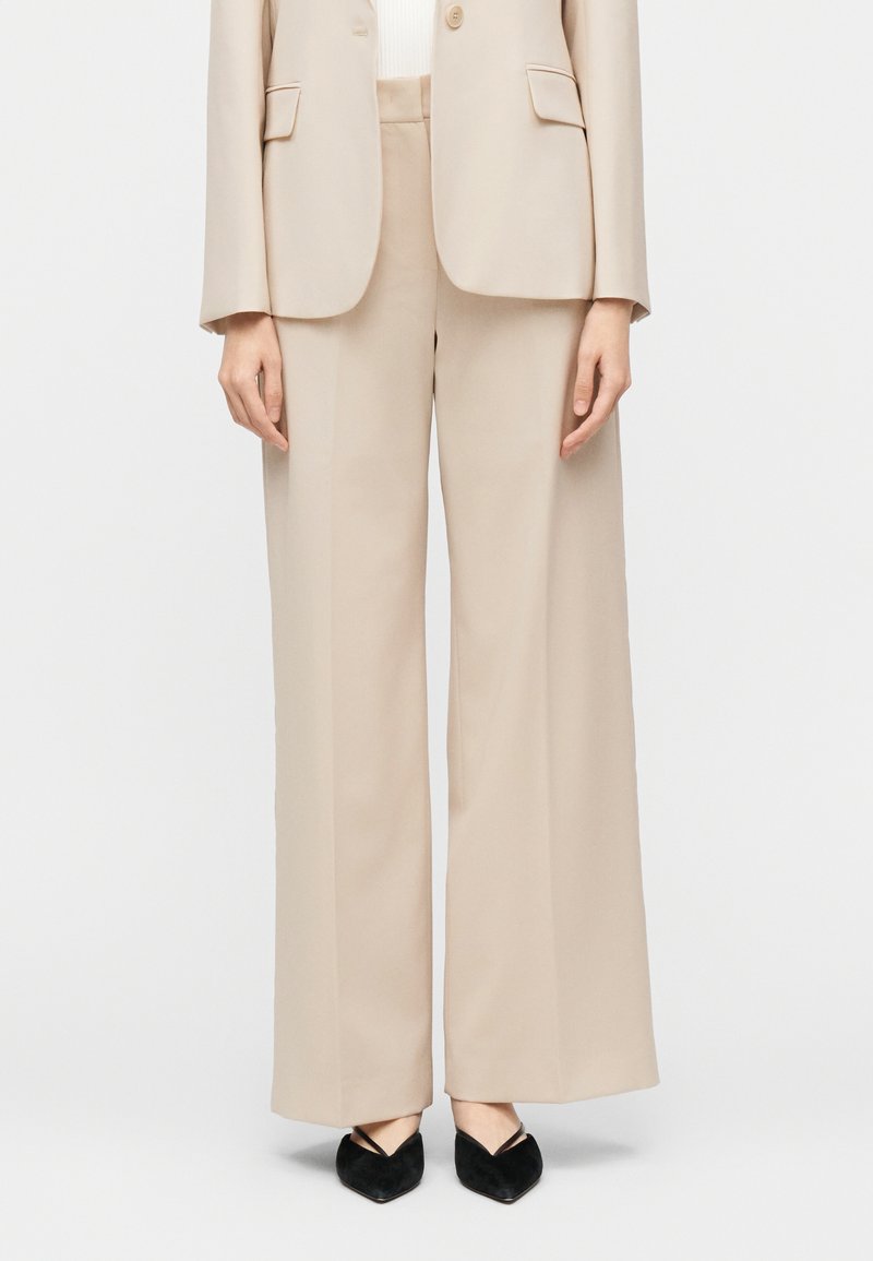 Μπεζ παντελόνια με φαρδιά μπάτζα ψηλή μέση, σετ με ταιριαστό μπεζ σακάκι και μαύρα παπούτσια με στρίπες και μυτερή μύτη.