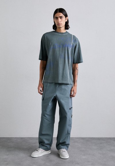 Camiseta gris de manga corta con texto azul, combinada con pantalones cargo grises de corte holgado que tienen bolsillos y detalles con cremallera, y zapatillas blancas.