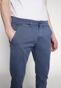 Tmavě modré bavlněné chino kalhoty s klasickým střihem, předními kapsami, zapínáním na knoflíky a jemnými šicími detaily.