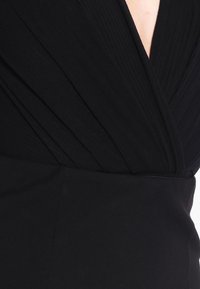 Robe noire avec un décolleté en V plongeant, présentant une texture de tissu froncé et des plis ; ceinture lisse en dessous. Aucun motif ou accent supplémentaire visible.