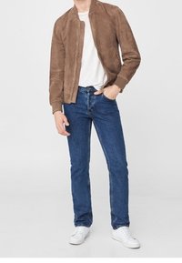 Man in een bruine suede jas, witte T-shirt, blauwe jeans en witte sneakers, staand met één hand in zijn zak tegen een egale achtergrond.