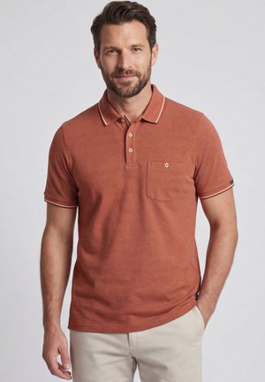 Man draagt een roestkleurig poloshirt met korte mouwen en zakje en beige broek, staand tegen een effen lichtgrijze achtergrond.