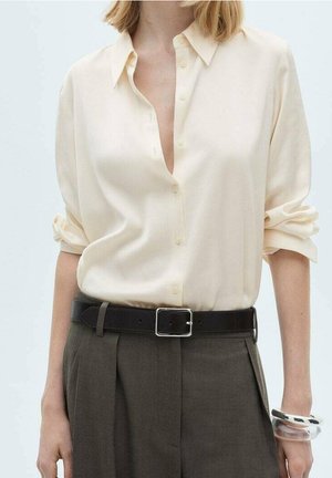 Femme portant une chemise crème boutonnée avec les manches retroussées, un pantalon sombre taille haute avec ceinture, et un bracelet clair et épais au poignet droit.