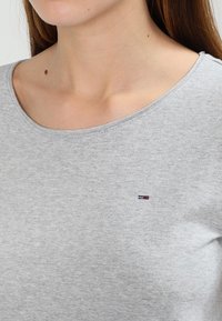 Grå bomullströja med rund halsringning och en liten broderad logotyp i röd, vit och blå på vänster bröstområde.