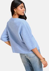 Jasnoniebieski sweter z krótkimi rękawami i luźnym krojem, o prążkowanej strukturze. Noszony z dżinsami o wysokim stanie i złotą bransoletką.
