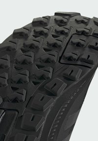 Semelle de chaussure de sport noire avec un design rugueux et texturé, dotée de crampons multidirectionnels pour une traction optimale et d'un bord lisse et durable.