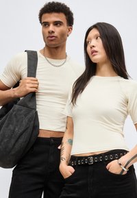 Άνδρας και γυναίκα φορούν μπεζ εφαρμοστά μπλουζάκια και μαύρο παντελόνι, ο άνδρας κρατάει μια μεγάλη μαύρη τσάντα, η γυναίκα έχει τατουάζ και ζώνη με μεταλλικά κορδόνια.