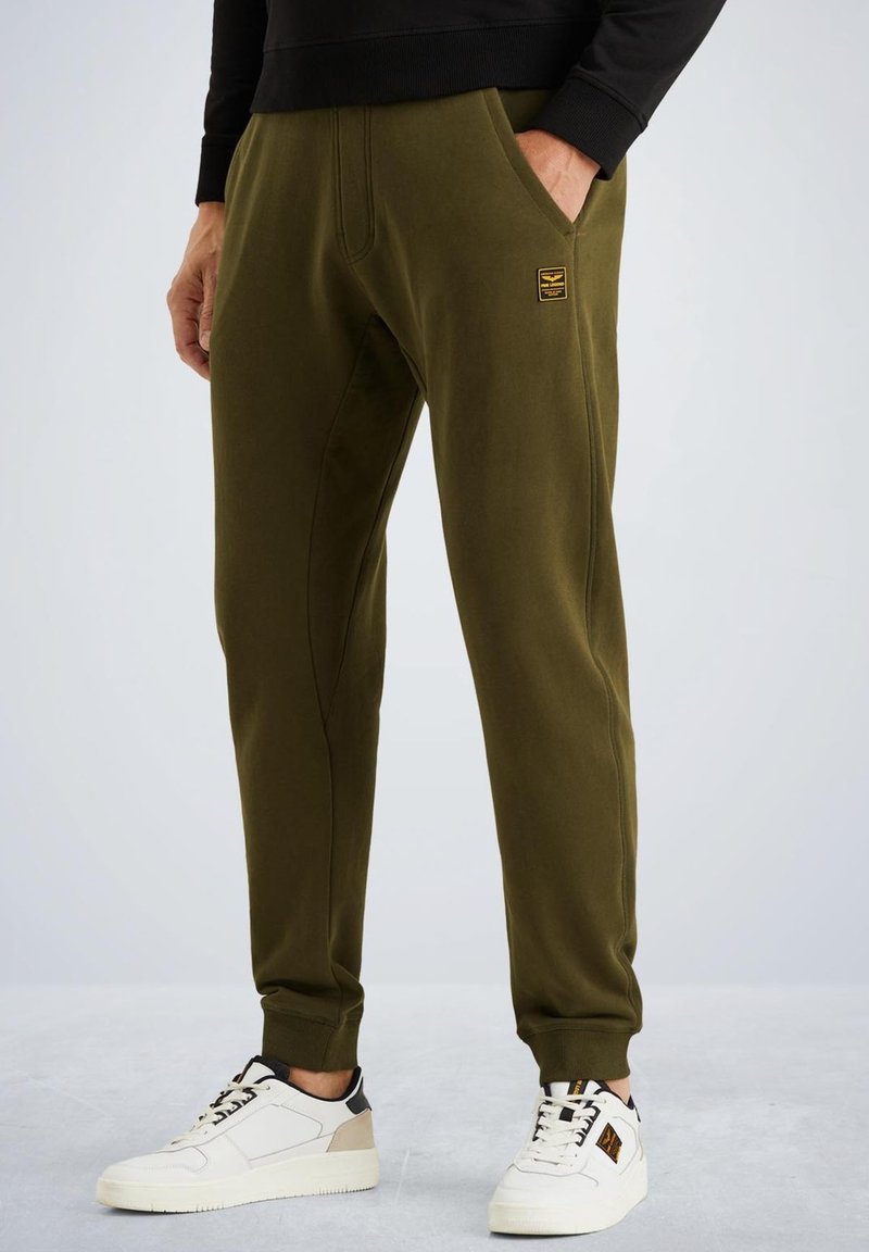 Olijfgroene joggingsbroek met een zachte textuur, voorzien van zijzakken en een zwart logo-embleem. Gekoeld met witte en zwarte sneakers.