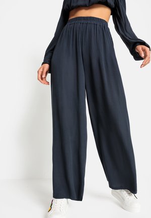 Hoge taille, brede broek in donkerblauw met een textuurafwerking. Benadrukt met een elastische tailleband voor comfort. Gecombineerd met witte sneakers.