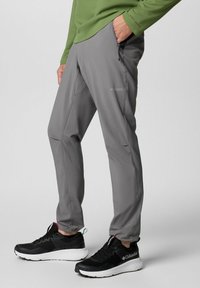 Pantalons de plein air légers et gris avec une coupe ajustée, dotés de poches zippées et de détails cousus. Associés à des chaussures de sport noires.