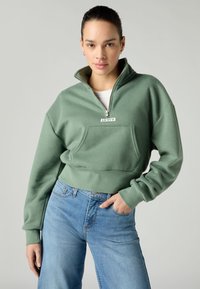 Sweat-shirt vert court avec un col zippé, une poche avant et le logo "LEVIS". Associé à un jean bleu clair. Texture lisse et coupe décontractée.