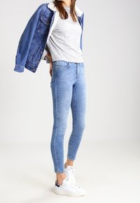Chaqueta de mezclilla con cuello de borreguito, camiseta gris, jeans ajustados de color azul claro y zapatillas blancas con acentos en azul marino. Conjunto sencillo y casual.
