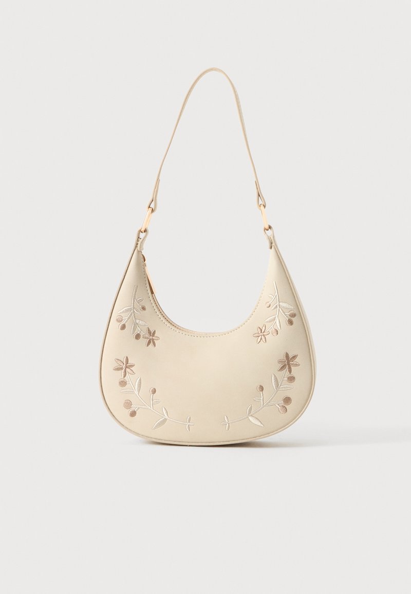 Sac à main en forme de croissant couleur crème avec une broderie florale et une courte sangle, suspendu sur un fond blanc.