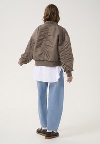 Brown satin bomber jacket with puffed sleeves, ribbed cuffs, and collar, over a white shirt, paired with light blue straight-leg jeans and dark shoes.