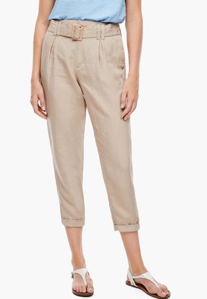 Vrouw draagt beige, hooggetailleerde, cropped broek met ceintuur en witte open sandalen, gecombineerd met een lichtblauw top, staand tegen een witte achtergrond.