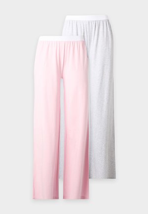 Kvinde står barfodet iført en hvid cropped tanktop og løse, lyserøde bukser mod en ensfarvet lys baggrund.