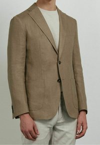 Blazer in lino color marrone chiaro con rever a lancia, due tasche frontali e chiusura con un solo bottone. Indossato sopra una camicia grigio chiaro.