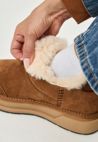 Mano che aggiusta un pantofole in suede beige con rivestimento morbido color crema, indossando un calzino bianco e jeans in denim blu.