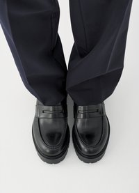 Svarte lærloafers med glatt finish, med en dekorativ stropp over vristen, kombinert med mørkeblå bukser.