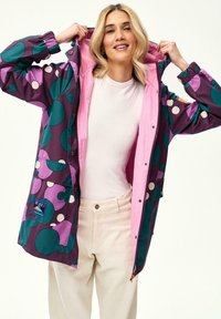 Kobieta w kolorowej kurtce z wzorem w odcieniach fioletu, zieleni i różu, z kapturem, białą bluzką i jasnobeżowymi spodniami, uśmiechająca się podczas podciągania kaptura.