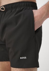 Sorte badebukser med snor i taljen, sidelomme og hvid "BOSS" logo på nederste kant. Glat stofstruktur.