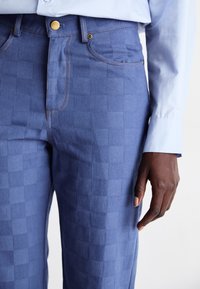 Prim-plan cu o persoană îmbrăcată în pantaloni cu carouri albastre și cusături portocalii, un nasture din alamă și un mânecă de cămașă lungă de culoare albastră deschisă vizibilă.