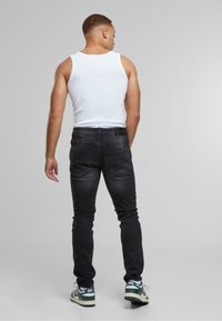 Man in een witte tanktop, zwarte jeans en groen-witte Nike sneakers, staand met zijn gezicht naar een effen lichtgrijze achtergrond.