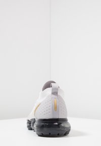 Achteraanzicht van een sneaker met een witte gebreide bovenkant, grijze accenten, gouden details en een zwarte doorzichtige zool met een gestructureerde hak.