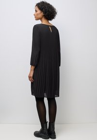 Schwarzes, plissiertes Kleid mit Dreiviertelärmeln, einem Schlüsselloch-Detail im Rücken und einem fließenden Design, kombiniert mit schwarzen Knöchelstiefeln.