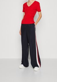 Person som bär en röd V-ringad kortärmad topp, marinblå vida byxor med vita och vinröda sidostrecker, samt vita plattformssneakers.