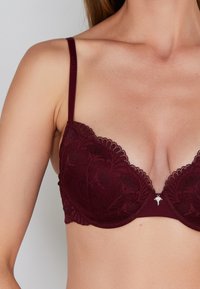 Burgundy lace bra with underwire, scalloped edges, and a center accent. Adjustable straps and smooth fabric with floral pattern detail.