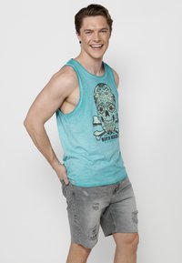 Turkos linne med en grafisk dödskalle-design och blommiga detaljer, i kombination med grå slitna denimshorts.