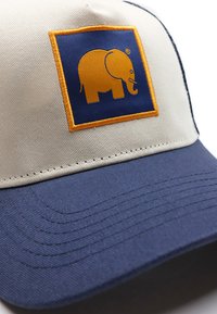 Vaaleanbeige ja tummansininen puuvillapallohattu, jossa on sininen neliön muotoinen kangas ja keltainen elefantti brodeerattuna. Litteä lippa ja teksturoitu kangas.