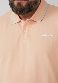 Pfirsichfarbendes Polohemd mit strukturiertem Gewebe, hellblauen Akzentstreifen am Kragen und einem weißen gestickten Logo auf der linken Brust.