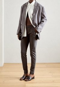 Person trägt grauen Blazer, weißes Hemd, schwarzes, bauchfreies Oberteil, schlanke graue Hose, schwarze Sandalen mit Absatz und hält eine kleine schwarze Clutch.