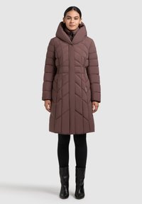 Sieviete, tērpusies garā brūnā ziemas virsjakā ar augstu apkakli, melnās legingās un melnās potītes zābakos, stāv pret pelēku fonu.