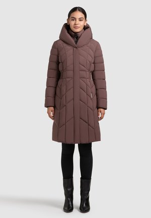 Nő hosszú barna steppelt télikabátban, magas gallérral, fekete leggingsben és fekete boka csizmában, szürke háttér előtt áll.